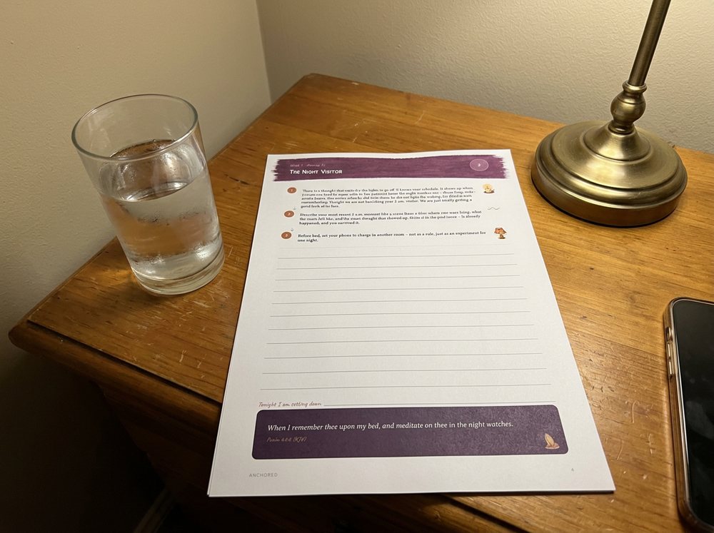 A printed evening page on a bedside table, under a lamp, beside a glass of water